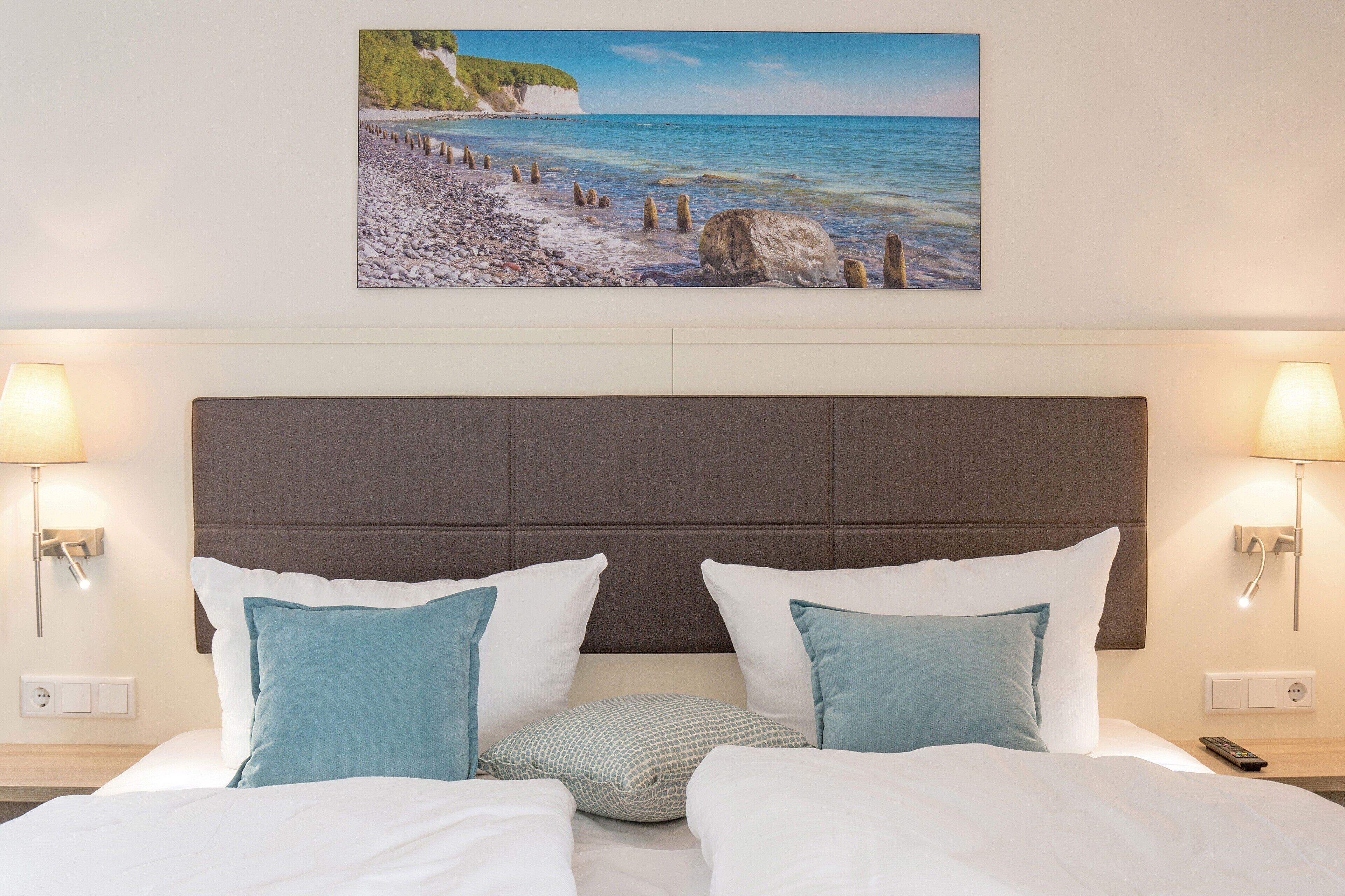 Une chambre d’hôtel avec un lit, deux lampes de chevet, une tête de lit et un tableau représentant une scène de plage.