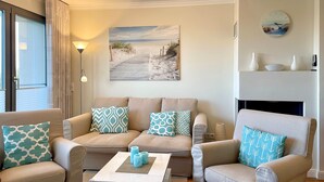 A living room with a beige sofa set, patterned cushions, a coffee table with decorative items, a painting of a beach pathway, and a clock on the wall. - Apartment 'Villa Deichgraf 07 Ostseeblick' with Sea View, Balcony and Wi-Fi (Börgerende-Rethwisch)