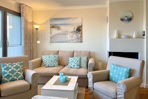 Ein Wohnzimmer mit einem beige-farbenen Sofagarnitur, gemusterten Kissen, einem Couchtisch mit Dekoration und einem Bild von einem Strandweg. An der Wand hängt eine Uhr.