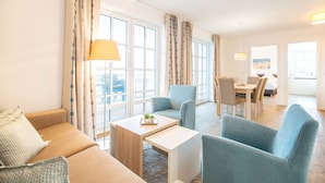 A modern hotel room with a sofa, armchairs, a dining table, and large windows. - Apartment 'Dünenrose 04 Wolkenlos' with Sea View, Balcony and Wi-Fi (Börgerende-Rethwisch)