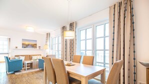 A modern living room with a sofa, coffee table, and dining table with chairs. - Apartment 'Dünenrose 04 Wolkenlos' with Sea View, Balcony and Wi-Fi (Börgerende-Rethwisch)