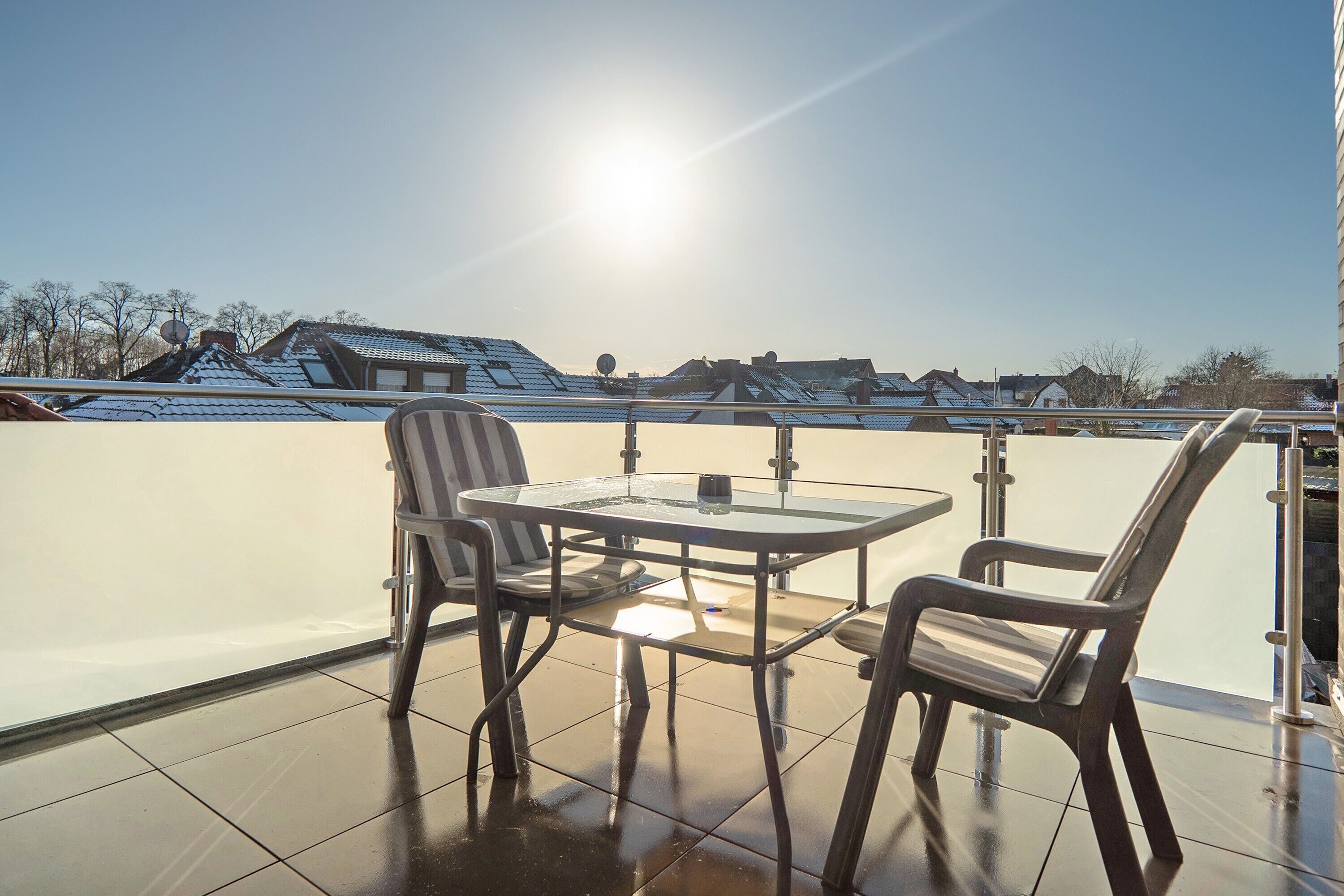 A balcony with a glass table and chairs, a clear view of the neighborhood, and a sunlit sky.