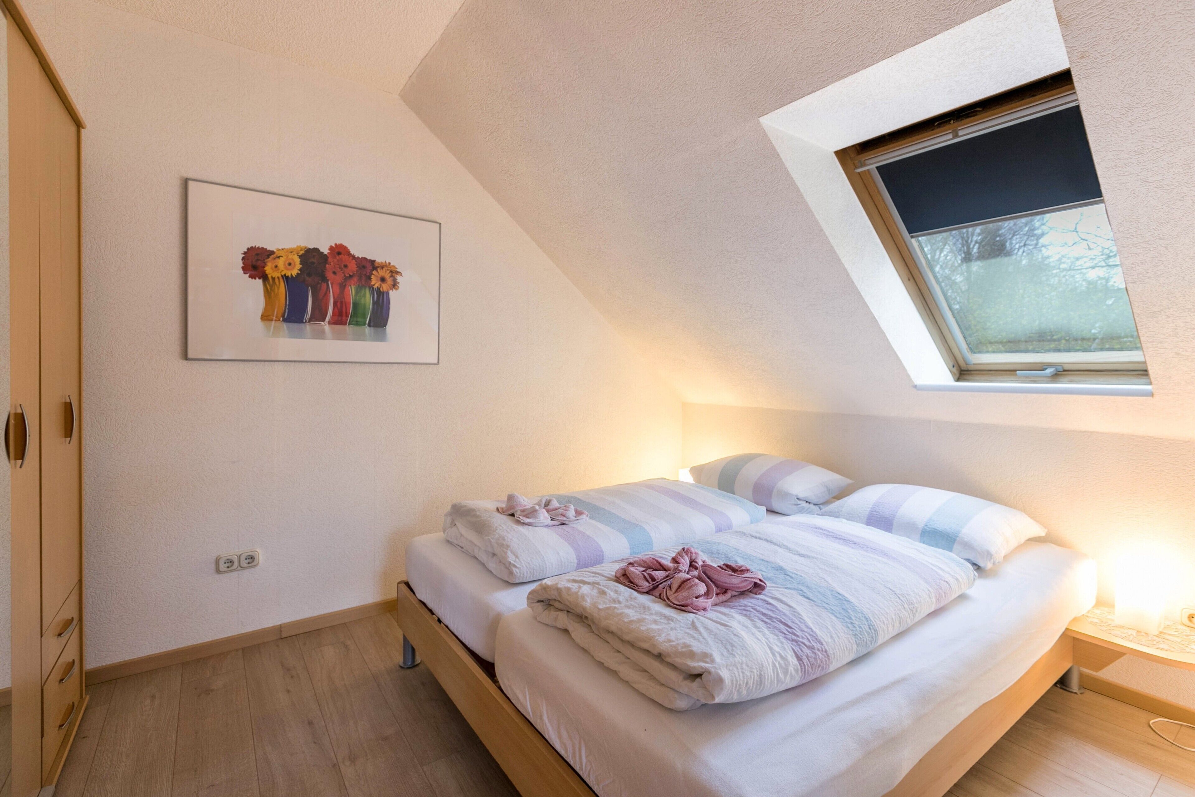 A bedroom with a skylight, a bed with striped bedding, a bedside table with a lamp, and a wall-mounted picture of vases.