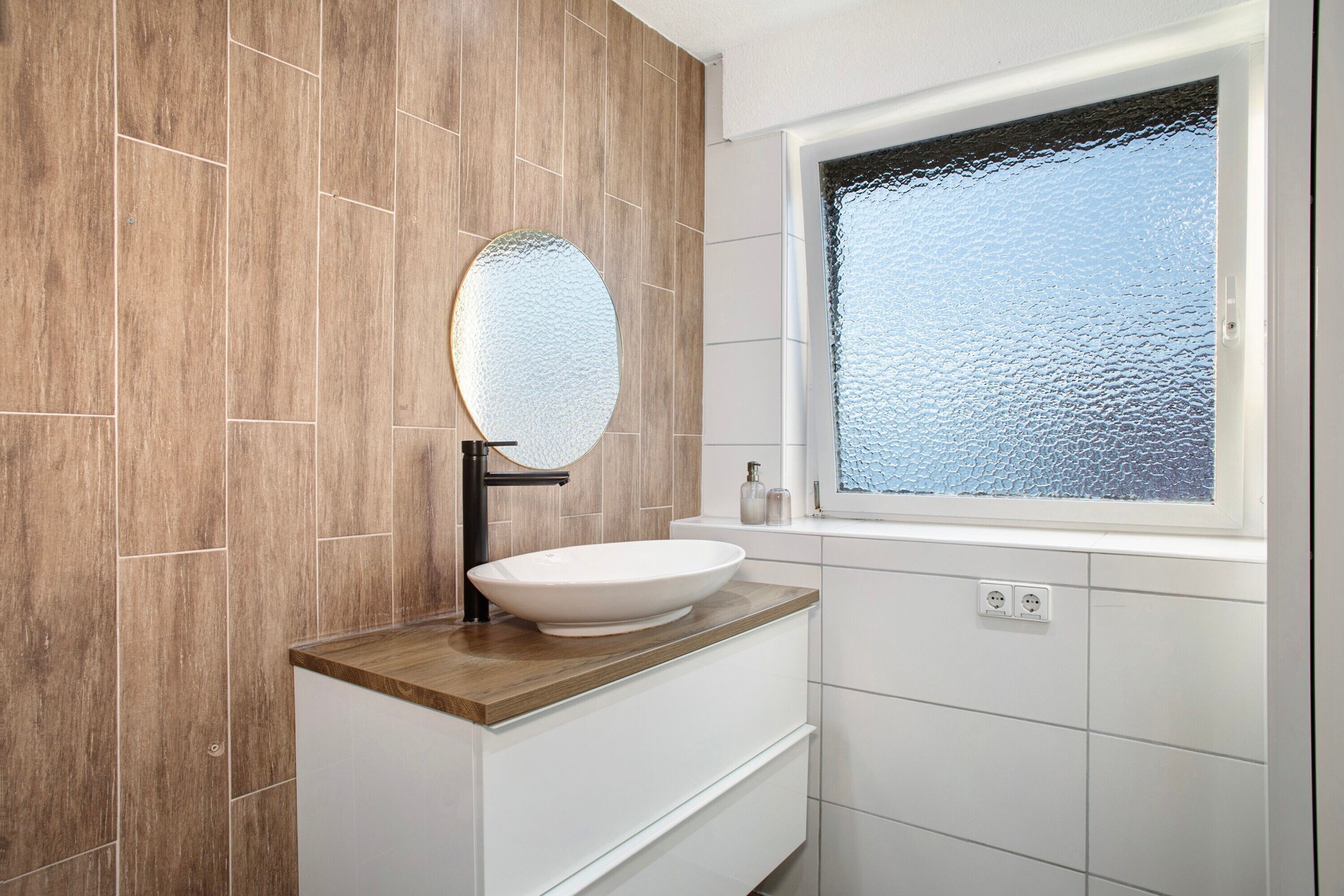 Un baño con pared de madera, un lavabo blanco con espejo redondo y una ventana con vidrio esmerilado.