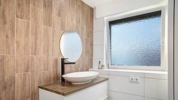 Un baño con pared de madera, un lavabo blanco con espejo redondo y una ventana con vidrio esmerilado.