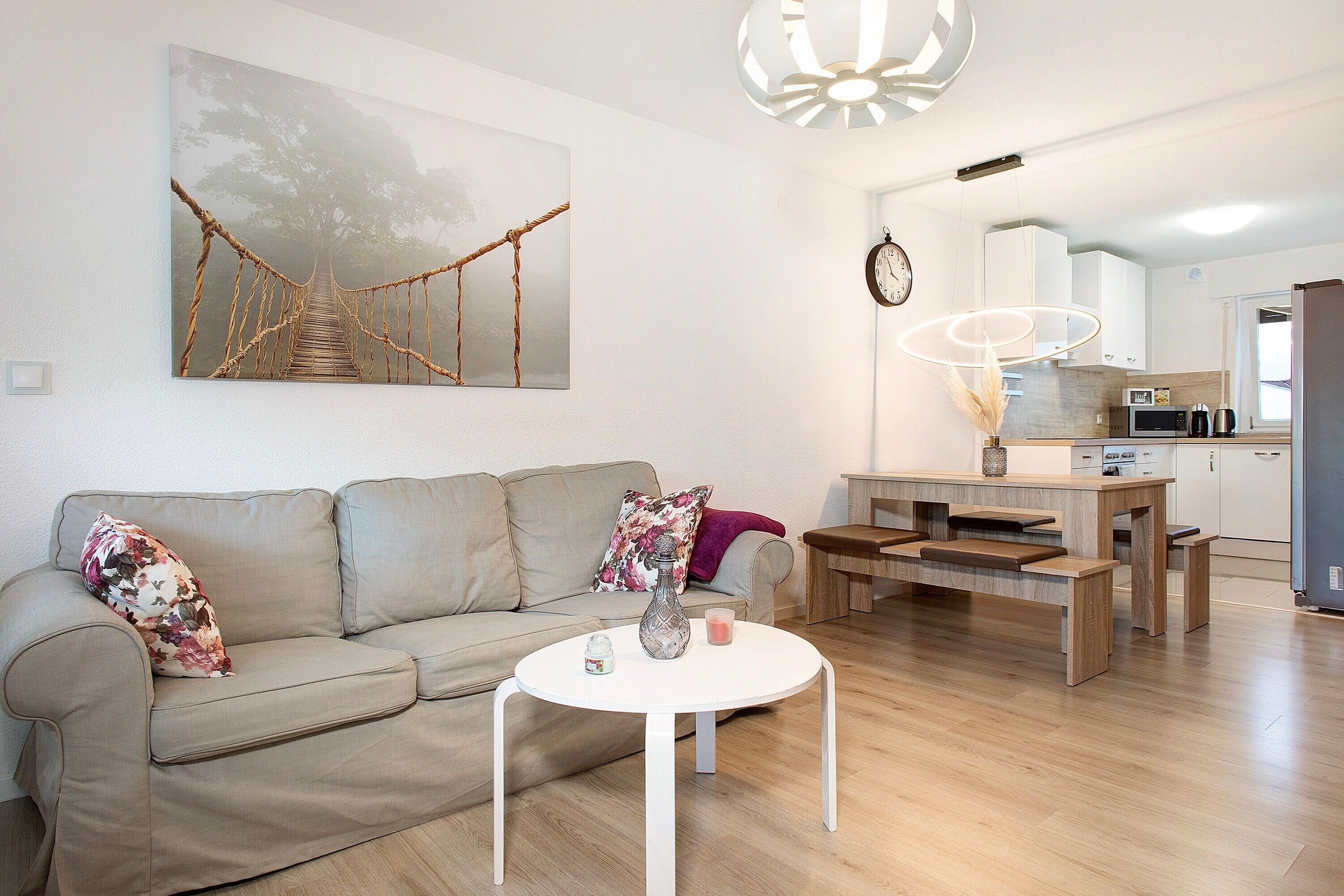 Una sala moderna con un sofá, una mesa redonda de centro, un cuadro de un puente, una zona de comedor con una mesa de madera y una cocina al fondo.