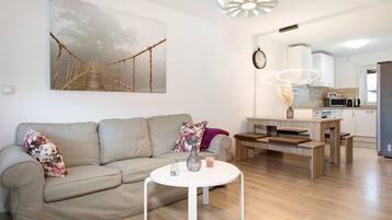 Una sala moderna con un sofá, una mesa redonda de centro, un cuadro de un puente, una zona de comedor con una mesa de madera y una cocina al fondo.