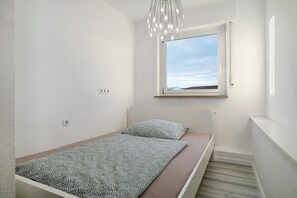 Una habitación pequeña con cama individual, ventana, lámpara en el techo y toma de corriente en la pared.