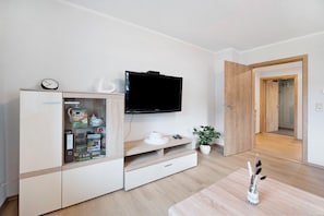 A modern living room with a TV, a cabinet with drawers, a small table with a vase, and a wooden door leading to another room.