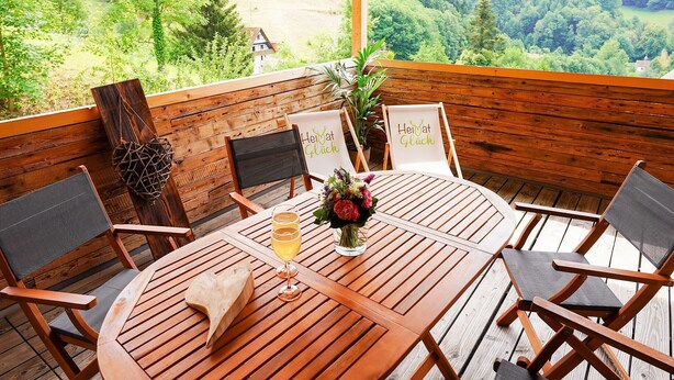 A wooden deck with a round table and chairs, a glass of wine, and a sign that reads 'Heimat Glück'. - Apartment 'Heimatglück 2, Maisonette, 76qm' with Shared Terrace, Balcony and Wi-Fi (Lautenbach)