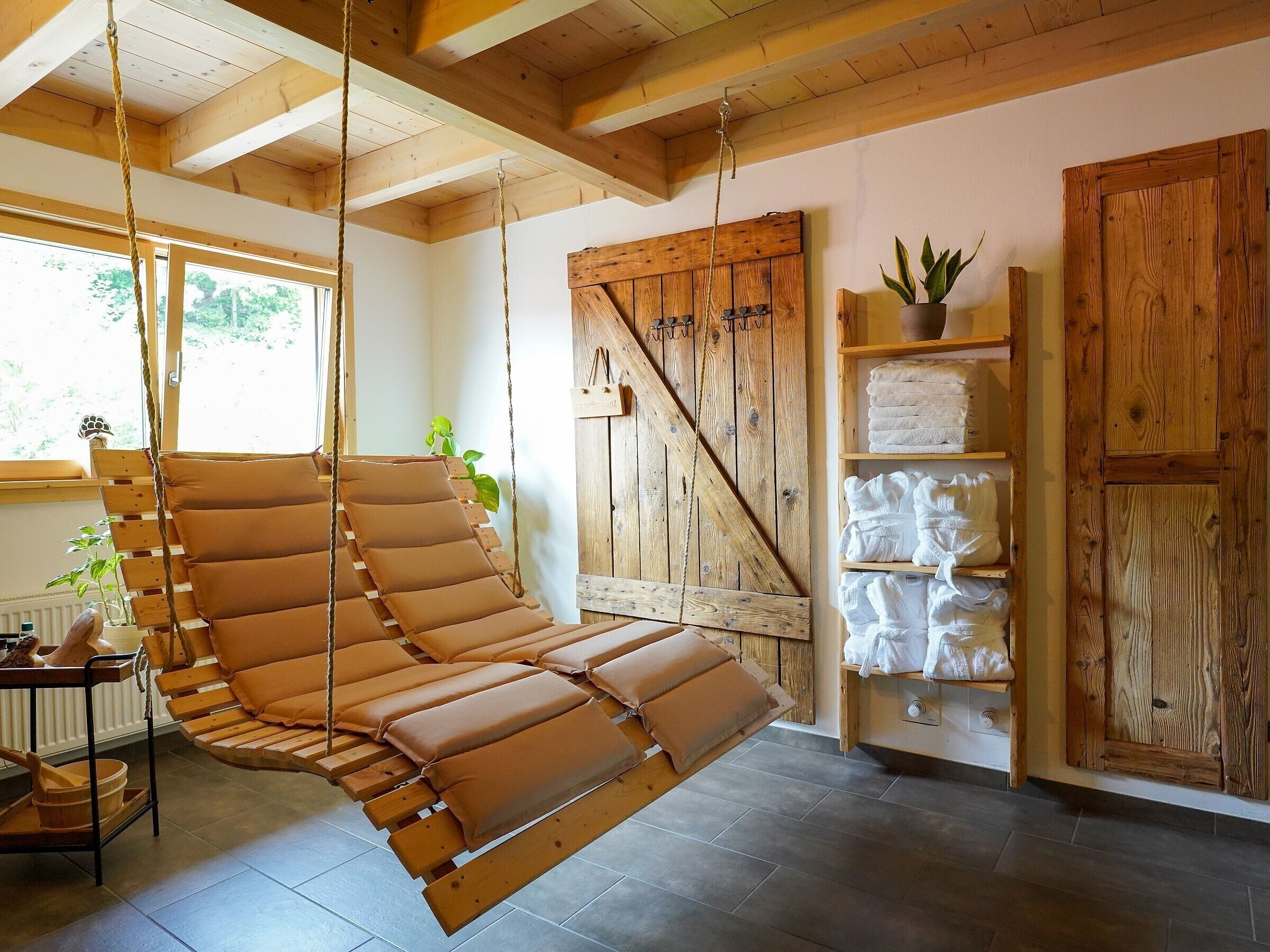 Een kamer met een hangstoel, houten planken met handdoeken, een houten deur en een raam met planten.