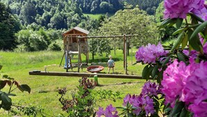 Una casetta giocattolo in legno con una scala e un’altalena in un’area erbosa con alberi e fiori.