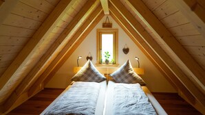 Una stanza con soffitto a falde, un letto con copriletto a quadretti, una finestra vista verde e una lampada fissata alla parete.