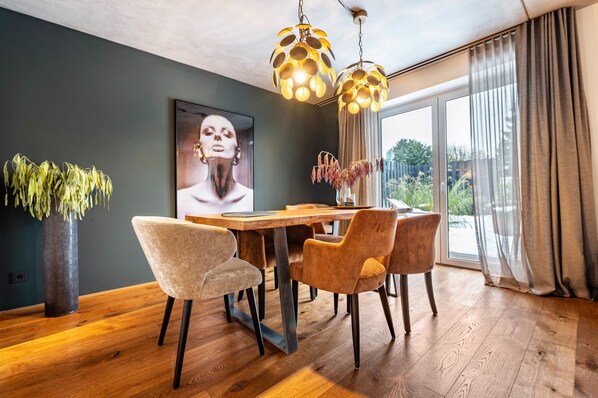 A dining area with a wooden table, four chairs, a vase with dried flowers, and a large window with curtains.