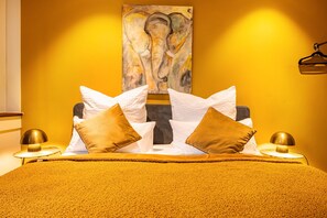 A bed with a yellow blanket, white and brown pillows, and a painting of an elephant on the wall.