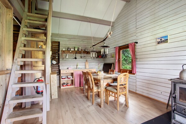 A wooden interior with a dining table, chairs, a wooden staircase, a stove, and a window with a view of greenery.
