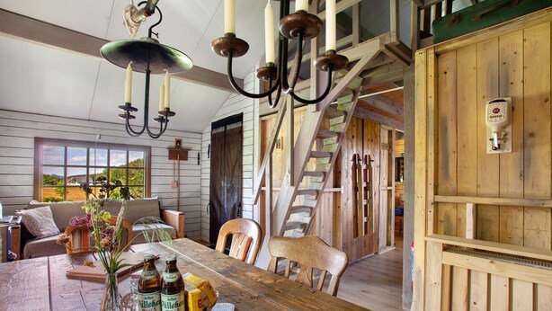 A wooden dining table with a vase of flowers, bottles, and a basket. A staircase leads to a loft area.