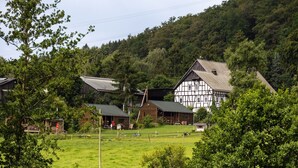 A traditional half-timbered house with a sloped roof, surrounded by a fenced pasture and dense forest.