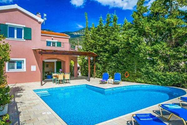 A swimming pool with blue water, surrounded by a pink building with green shutters, and a covered outdoor area with chairs and tables.