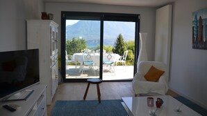 Ein Zimmer mit Fernseher, einem weißen Schrank, einem weißen Sessel mit orangefarbenem Kissen, einem kleinen Hocker und einem Tisch mit Vase und Kerze.