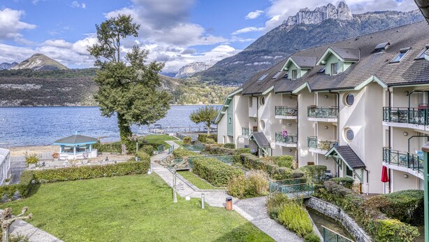 A hotel with multiple balconies, a garden area, and a view of a lake and mountains. - La plage privée du lac (Duingt)