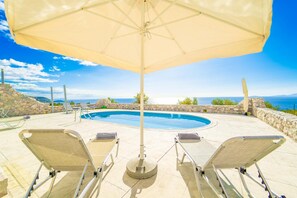 A poolside area with lounge chairs, an umbrella, and a view of the sea.