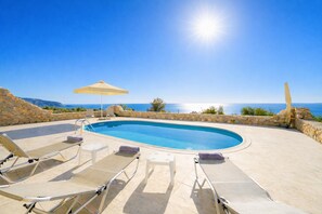 A swimming pool with lounge chairs and an umbrella, surrounded by stone walls and a view of the sea.