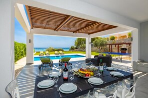 An outdoor dining area with a view of a pool and ocean, a table set for a meal, and a barbecue grill. - Villa Dionysos in Rhodes (Rhodes)