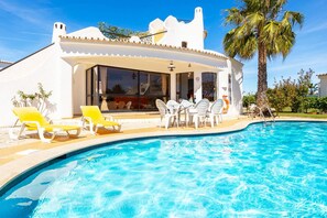 A poolside area with a white building, yellow and white lounge chairs, and a palm tree.