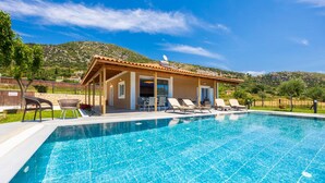 Piscina con sillas de descanso, un porche cubierto y montañas al fondo.
