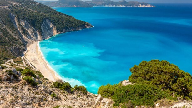 Un paisaje costero con mar azul transparente, una playa arenosa y colinas verdes.