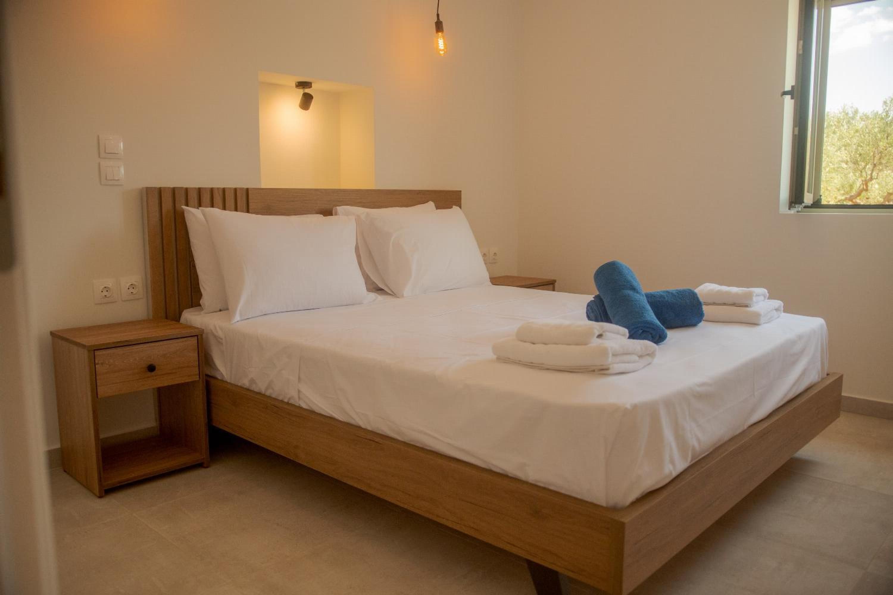 Een net opgemaakt bed met witte lakens en blauwe handdoeken, een houten hoofdbord, een nachtkastje met lade en een wandlamp.