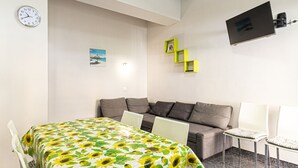 Une pièce moderne avec un canapé, une table recouverte d’une nappe à motifs de tournesols, un téléviseur fixé au mur et un petit cadre photo.