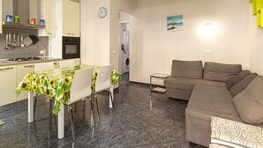 Une cuisine moderne avec des meubles blancs, une table à manger recouverte d’une nappe à motifs de tournesols et un canapé gris dans l’espace salon.