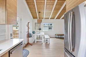 A modern kitchen with a stainless steel refrigerator, wooden ceiling beams, a dining area with a white table and bench, and a view through a window.