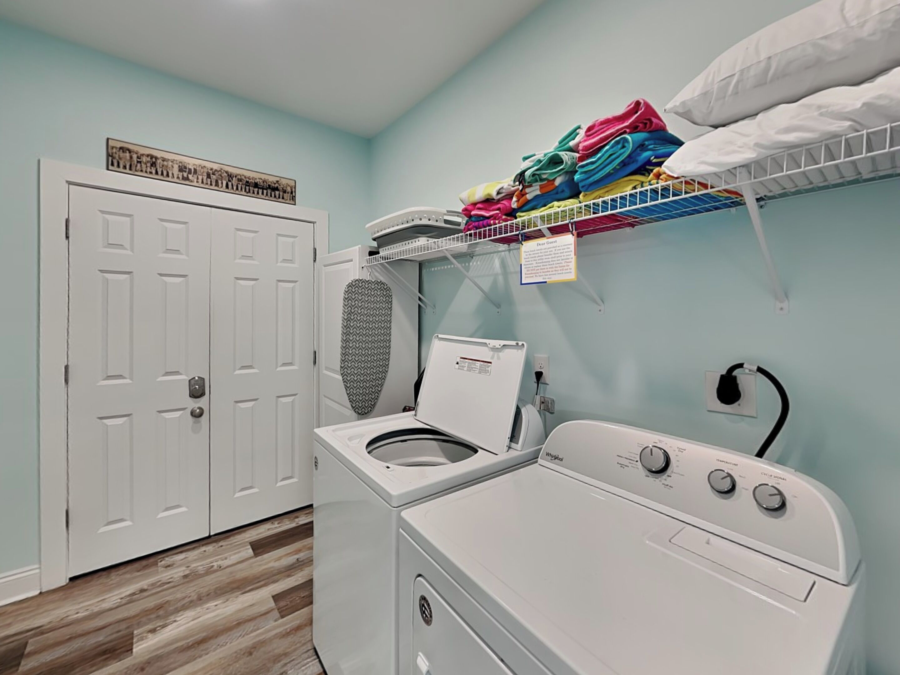 A laundry room with a front-loading washer and dryer, a drying rack with folded clothes, a ceiling-mounted fan, and a door with a framed picture.