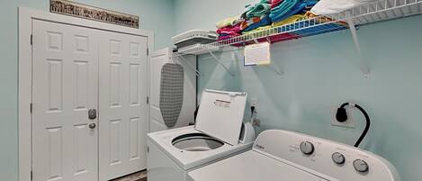 A laundry room with a front-loading washer and dryer, a drying rack with folded clothes, a ceiling-mounted fan, and a door with a framed picture.