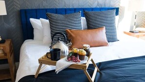 Een netjes opgemaakt bed met een blauw hoofdeinde, een dienblad met een theepot, een kopje en muffins, en een bruin kussen op een witte sprei.