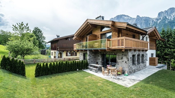 A modern two-story house with a stone facade, wooden balconies, and a view of mountains.