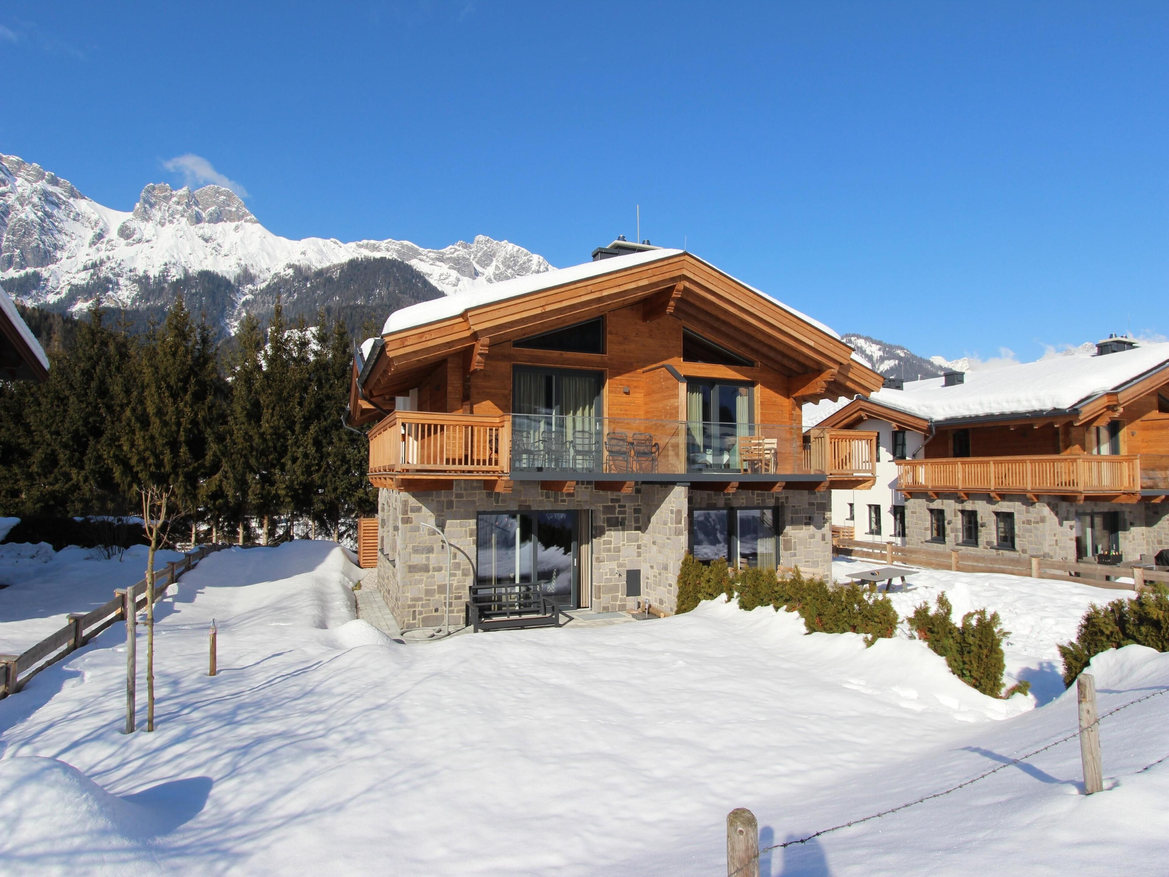 A wooden chalet with a balcony, stone foundation, and a view of snow-covered mountains.