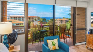 A room with a large sliding glass door leading to a balcony with a view of the ocean and buildings. There is a blue armchair with a pineapple pillow, a table with chairs, and a fan on a nightstand. - Top Floor Maui Vista with Ocean View, AC, 5min Walk to Beach (Kihei)