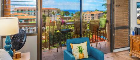 A room with a large sliding glass door leading to a balcony with a view of the ocean and buildings. There is a blue armchair with a pineapple pillow, a table with chairs, and a fan on a nightstand.
