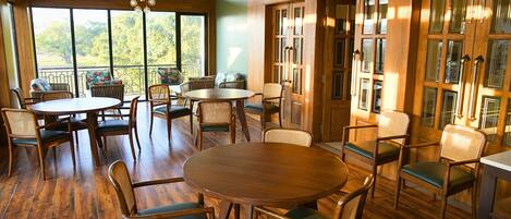 A spacious dining area with wooden tables and chairs, large windows offering a view of greenery, and a balcony with wicker furniture.