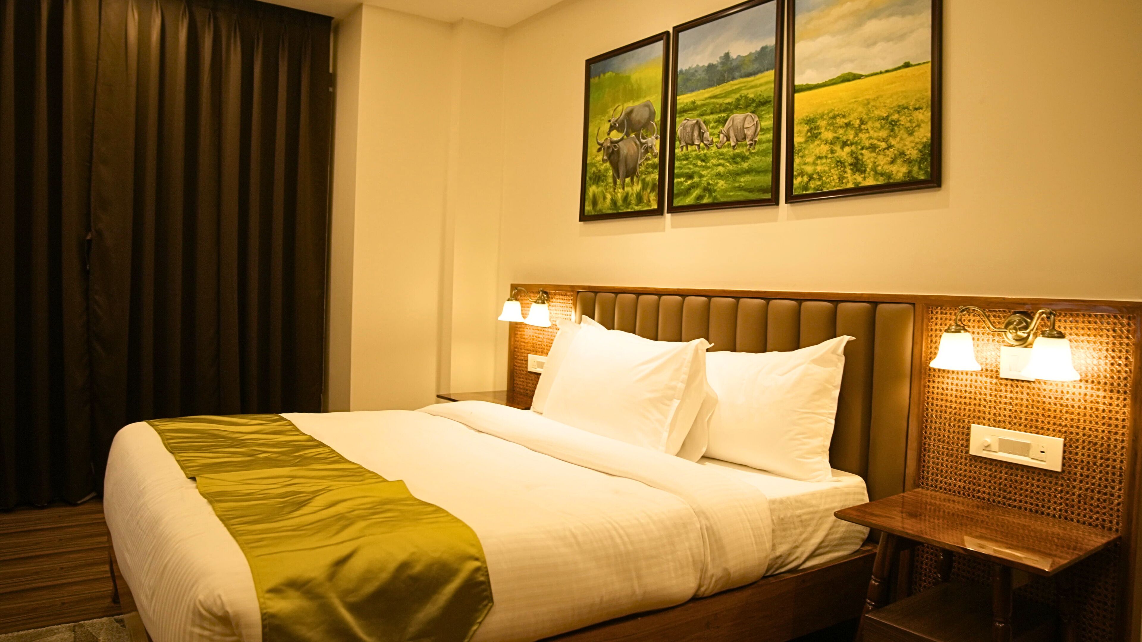 A hotel room with a large bed, two bedside lamps, a headboard with a textured pattern, and three framed paintings on the wall.