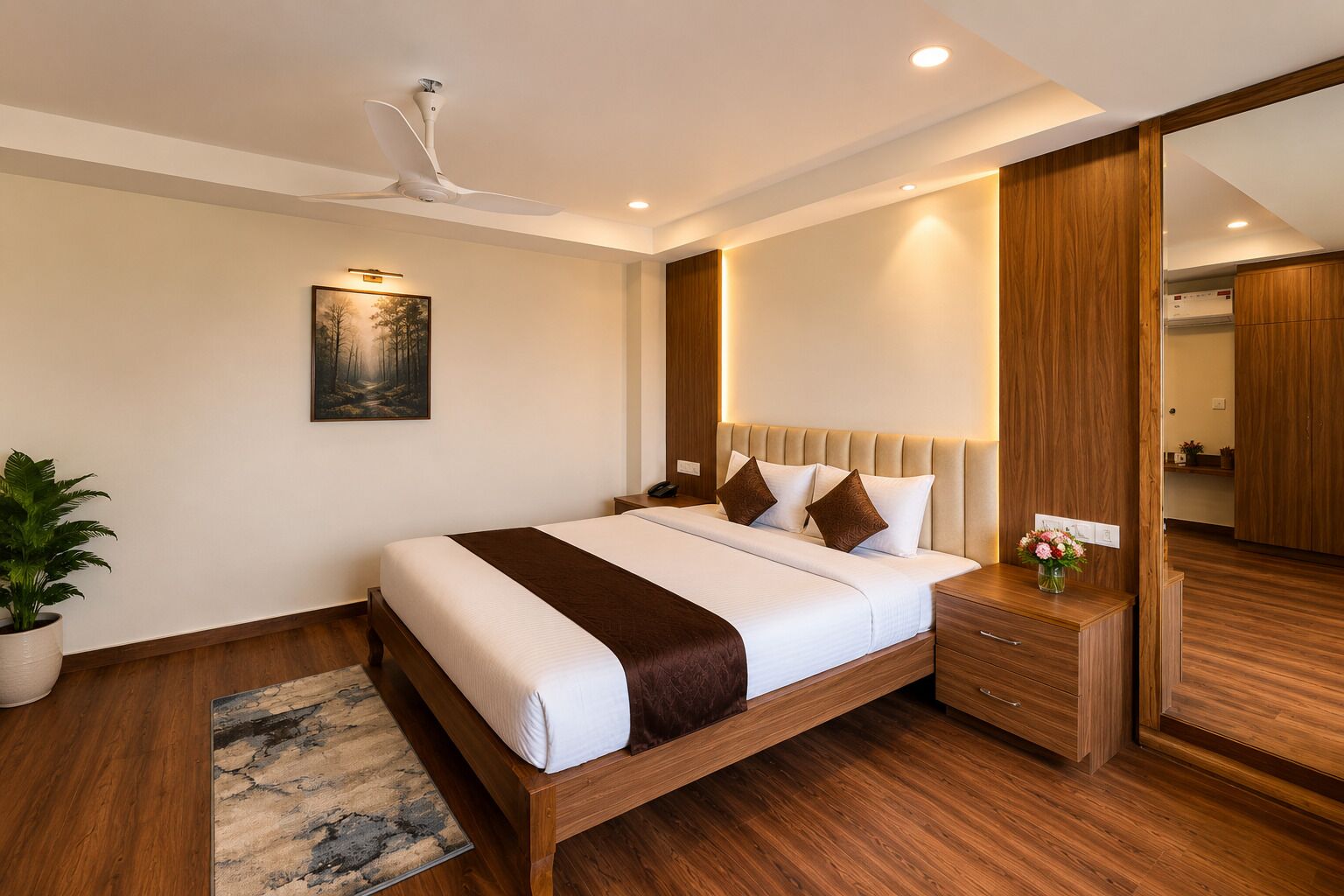 A modern bedroom with a wooden bed, a ceiling fan, a nightstand, a bedside table with a vase, and a large mirror.