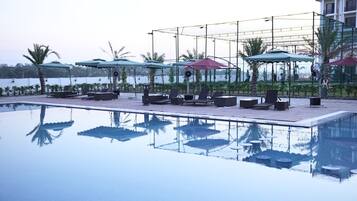 Piscina con sillas de descanso y sombrillas, rodeada de jardines con palmeras y vistas a un cuerpo de agua.