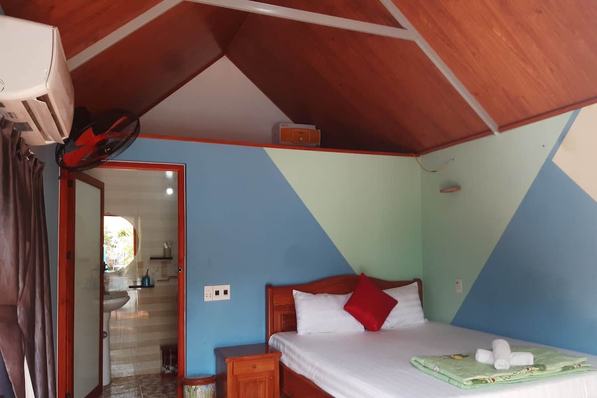 A bedroom with a wooden ceiling, a bed with white and red bedding, a small bedside table, and a green mat on the bed.