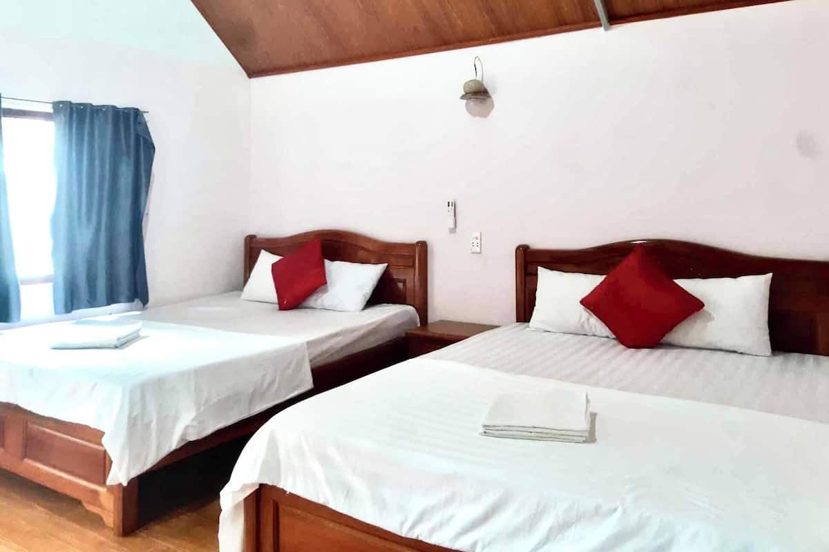 Two wooden beds with white linens and red pillows, a wooden floor, and a window with blue curtains.