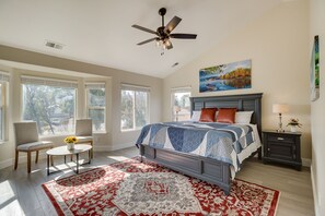 Un dormitorio con cama, dos sillas, una mesa con flores, un cuadro y una mesita de noche con lámpara.