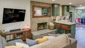Ein modernes Wohnzimmer mit Kamin, einem an der Wand befestigten Fernseher und einer Küchenzeile mit grünen Schränken und einer Bar.